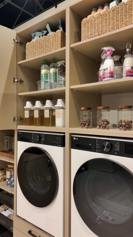 Open planken in wasmachinekast met wasmiddelen, potten en schoonmaakproducten naast twee LG wasmachines